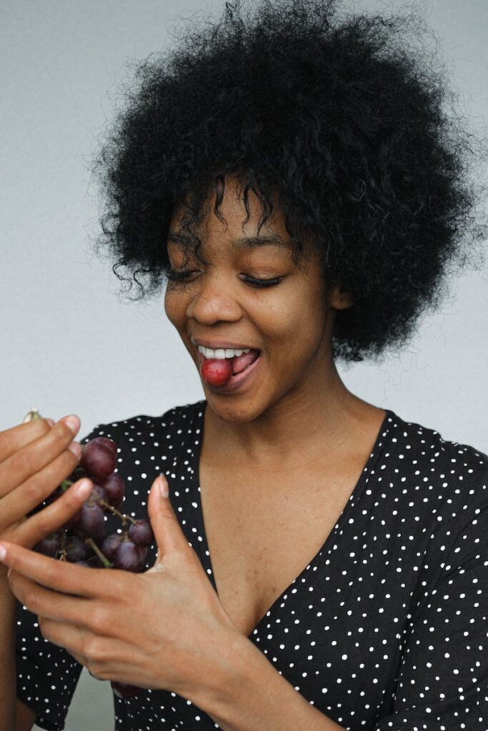 Young African American woman in polka dot dress trying delicious sweet grape on gray background